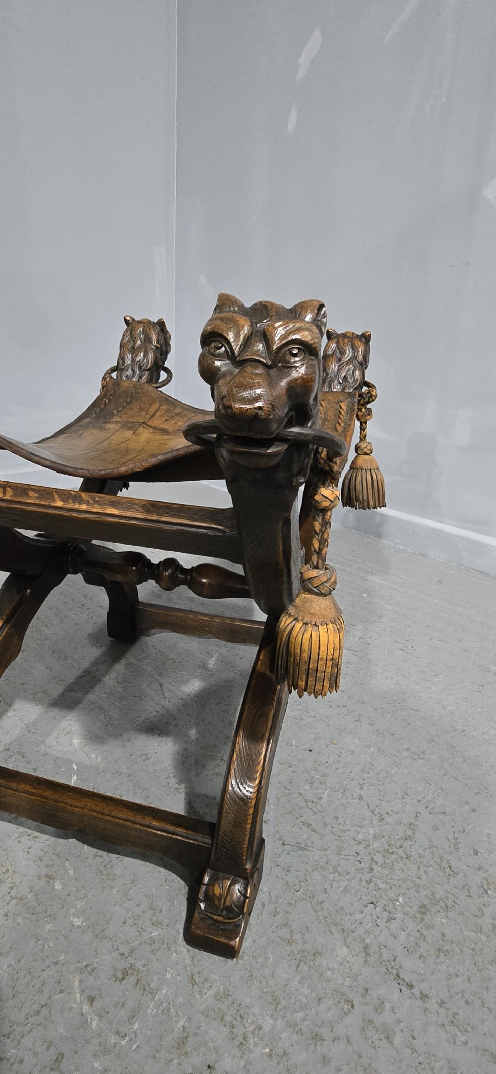 A carved dark walnut X-frame curule stool featuring lion heads with carved wooden tassels at the top corners and a slung brown leather seat. View VIII