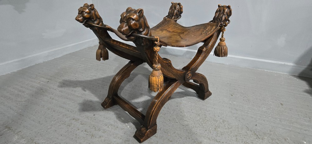 A carved dark walnut X-frame curule stool featuring lion heads with carved wooden tassels at the top corners and a slung brown leather seat. View XI