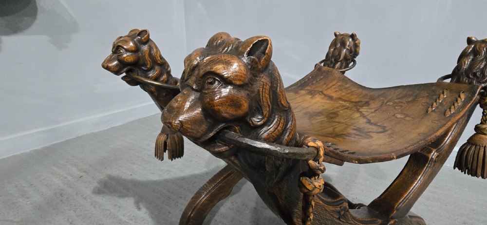 A carved dark walnut X-frame curule stool featuring lion heads with carved wooden tassels at the top corners and a slung brown leather seat. View XII