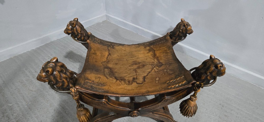 A carved dark walnut X-frame curule stool featuring lion heads with carved wooden tassels at the top corners and a slung brown leather seat. View XIII