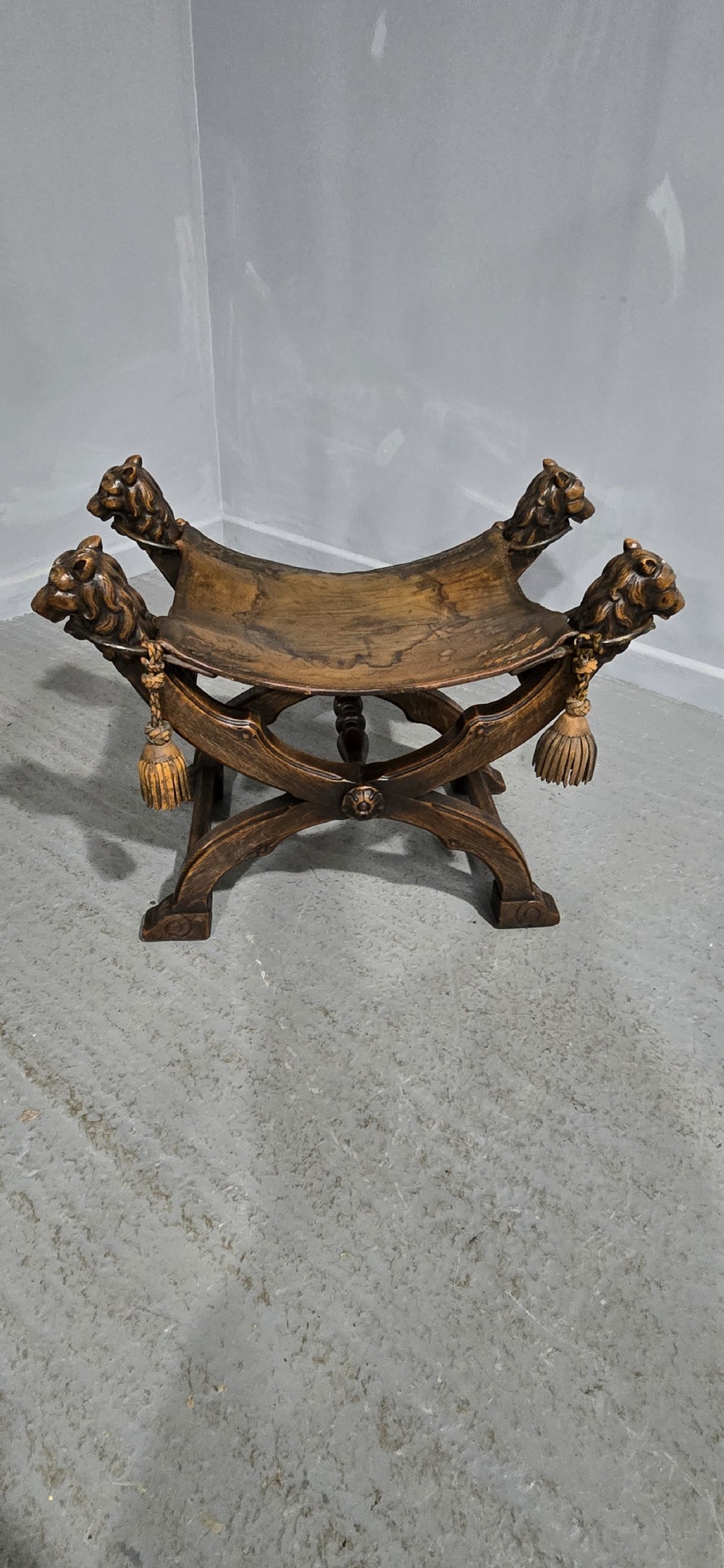 A carved dark walnut X-frame curule stool featuring lion heads with carved wooden tassels at the top corners and a slung brown leather seat. View XIV
