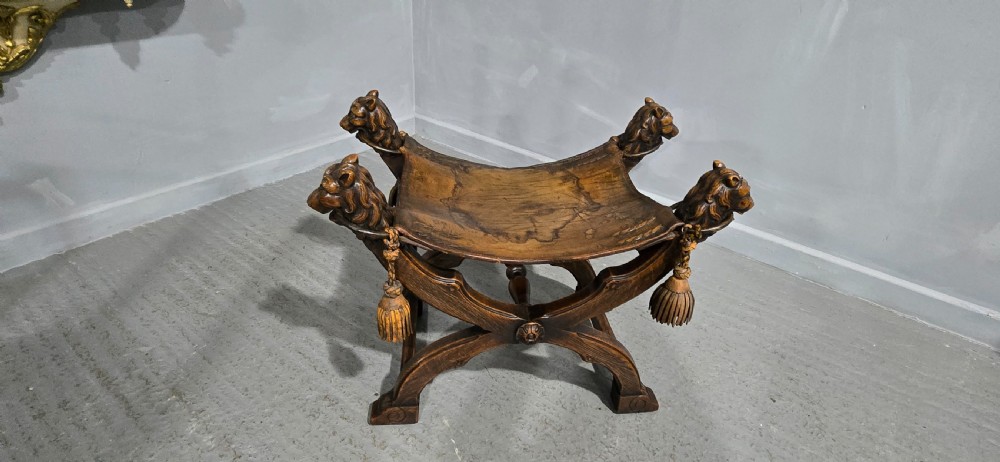 A carved dark walnut X-frame curule stool featuring lion heads with carved wooden tassels at the top corners and a slung brown leather seat. View XV