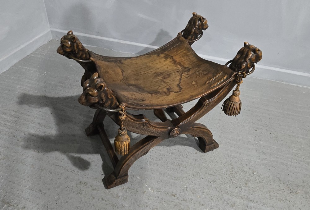 A carved dark walnut X-frame curule stool featuring lion heads with carved wooden tassels at the top corners and a slung brown leather seat. View XVI