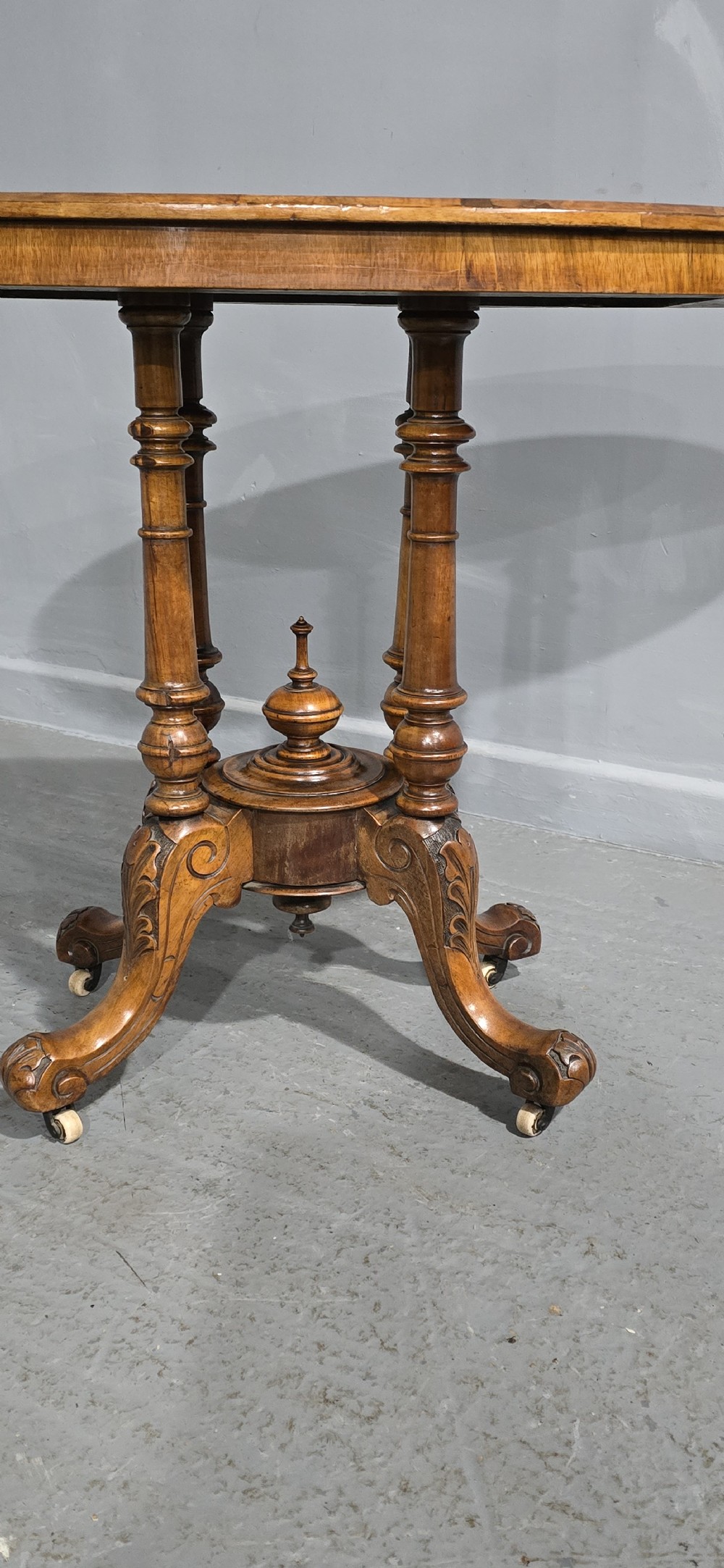 An oval Victorian centre table made from burr walnut with floral marquetry inlay, four turned support columns, and carved legs on castors. View II