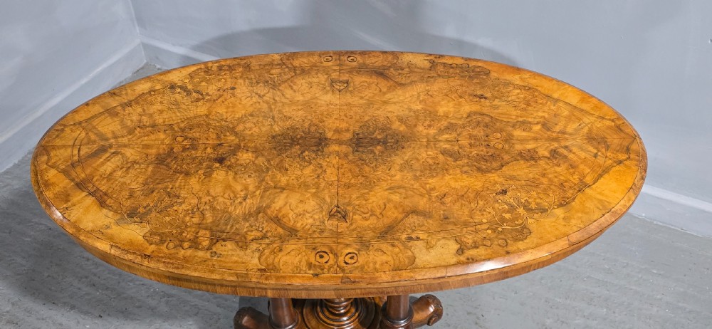 An oval Victorian centre table made from burr walnut with floral marquetry inlay, four turned support columns, and carved legs on castors. View VI