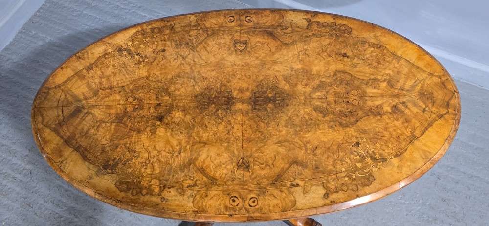 An oval Victorian centre table made from burr walnut with floral marquetry inlay, four turned support columns, and carved legs on castors. View IX