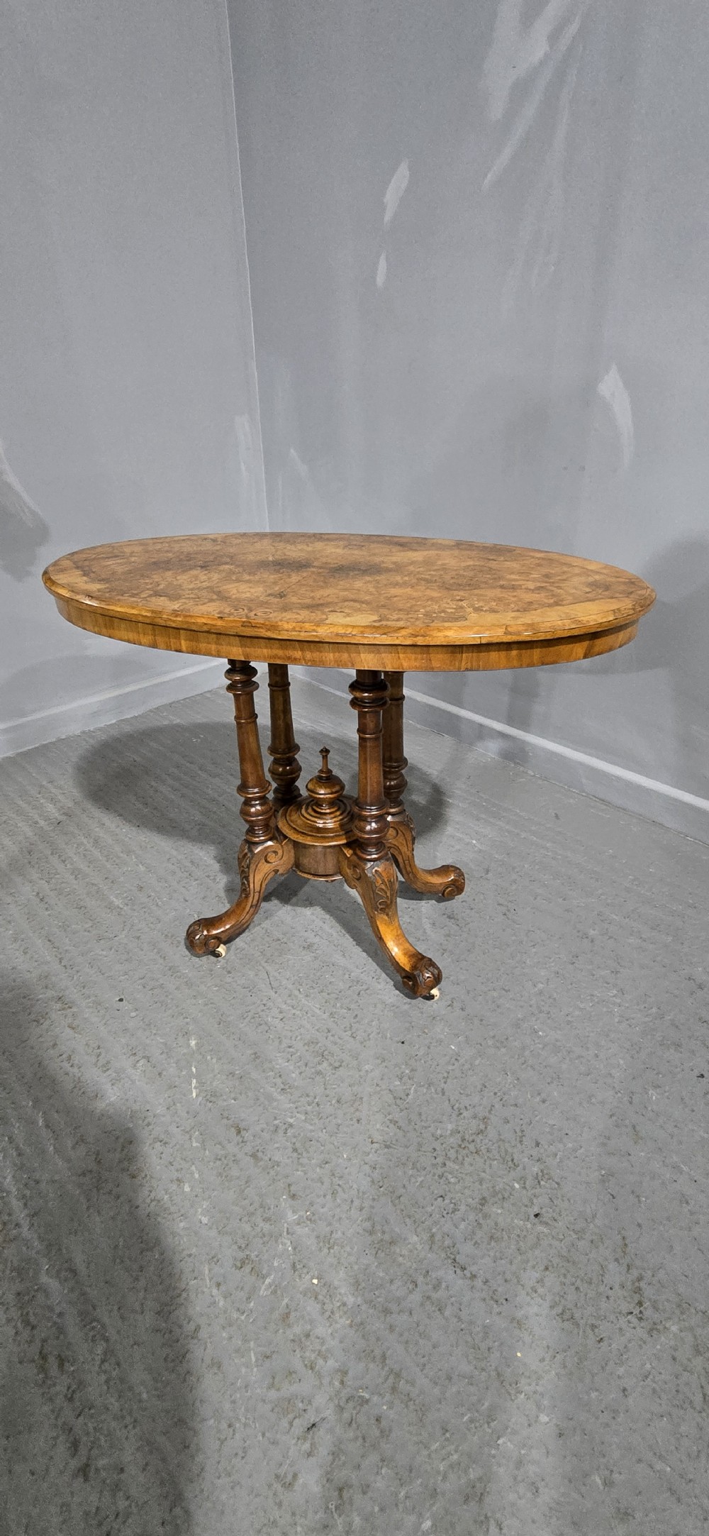 An oval Victorian centre table made from burr walnut with floral marquetry inlay, four turned support columns, and carved legs on castors. View XI