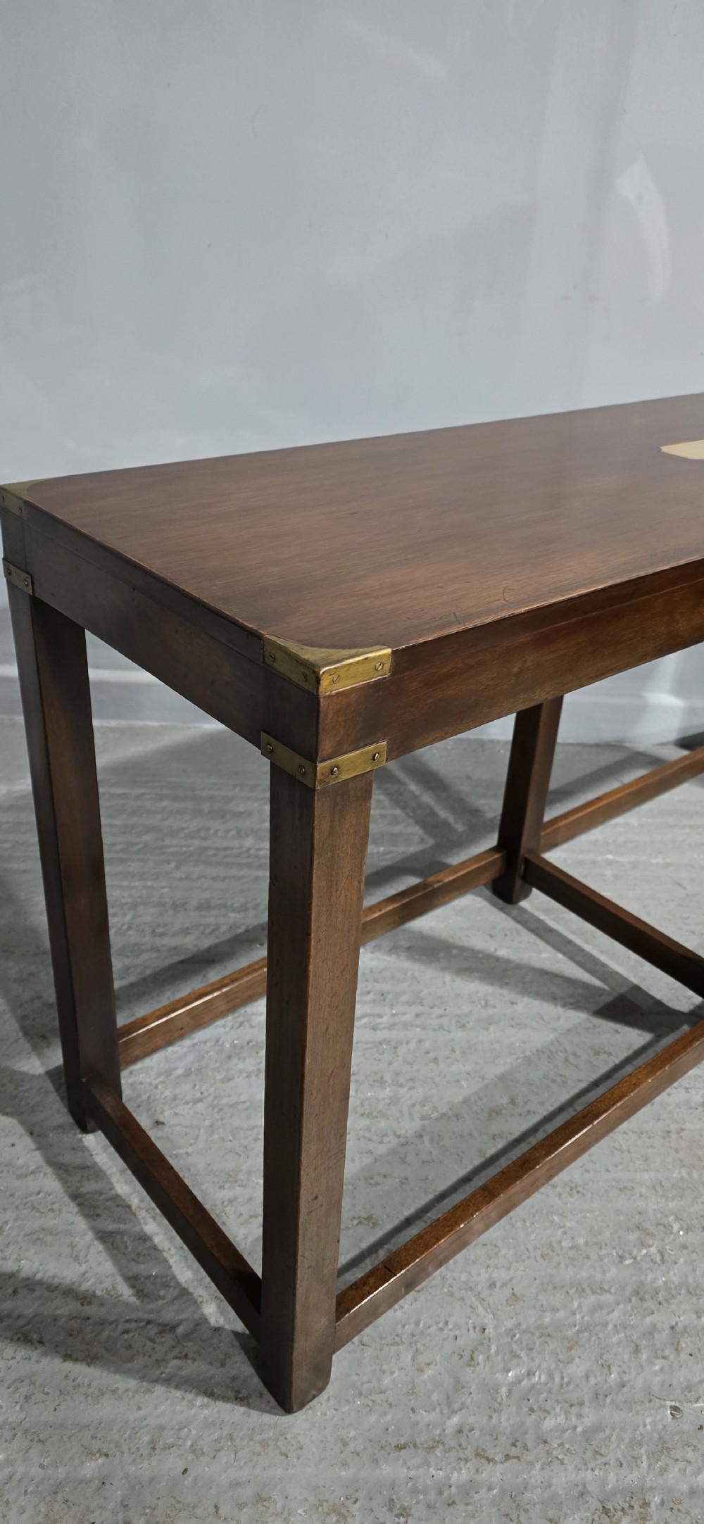 A rectangular mahogany campaign console table featuring brass corner mounts, an inset central brass plaque, and straight squared legs connected by a full box stretcher. View IV