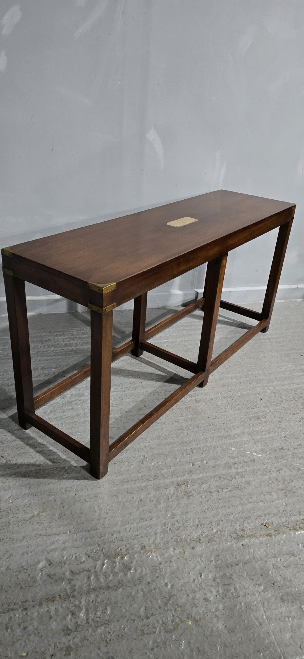 A rectangular mahogany campaign console table featuring brass corner mounts, an inset central brass plaque, and straight squared legs connected by a full box stretcher. View V