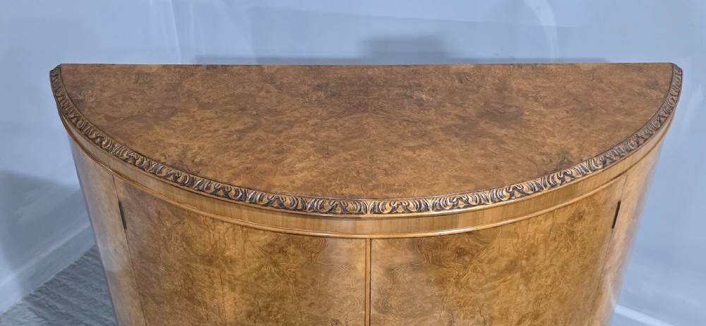 A semicircular burr walnut demi lune cabinet featuring double doors, figured wood grain, a central brass escutcheon with tassel, and carved cabriole legs. View VI