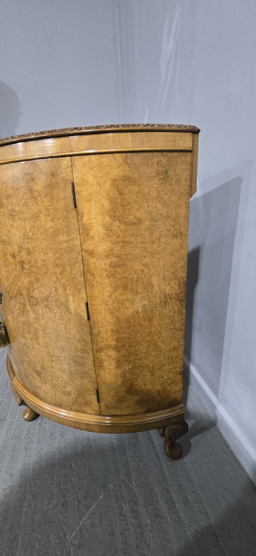 A semicircular burr walnut demi lune cabinet featuring double doors, figured wood grain, a central brass escutcheon with tassel, and carved cabriole legs. View XI