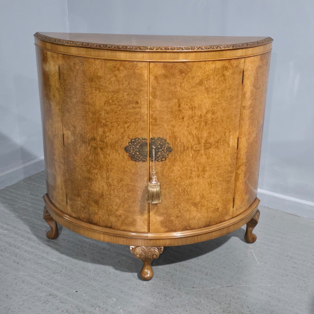 A semicircular burr walnut demi lune cabinet featuring double doors, figured wood grain, a central brass escutcheon with tassel, and carved cabriole legs.