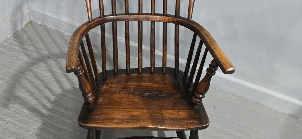 An antique dark brown timber Windsor armchair featuring a high hoop spindle back, curved armrests with turned supports, and a shaped solid wooden seat. View V