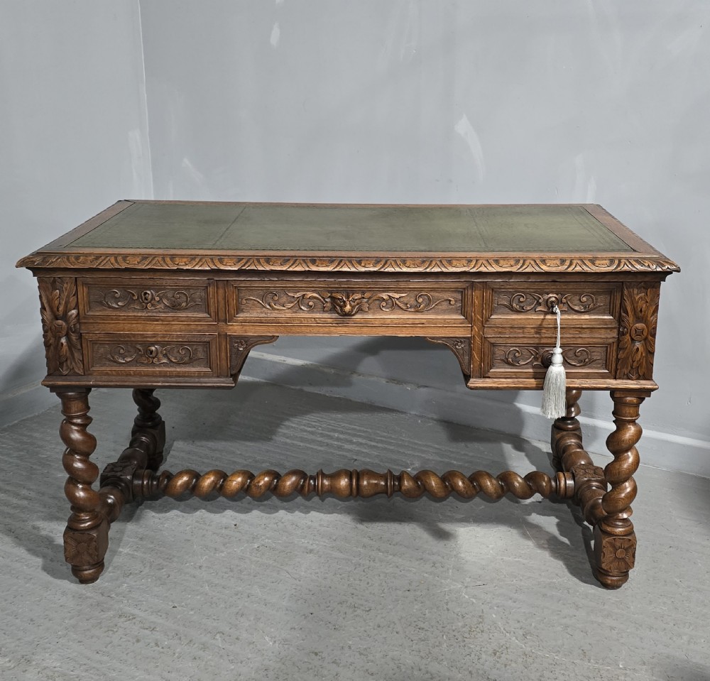 A carved oak writing desk featuring a green leather top insert, detailed scrollwork drawers with a central mask motif, and heavy barley-twist legs connected by a low stretcher.