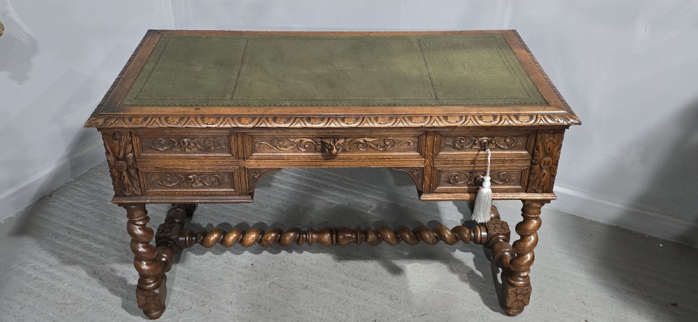 A carved oak writing desk featuring a green leather top insert, detailed scrollwork drawers with a central mask motif, and heavy barley-twist legs connected by a low stretcher. View II