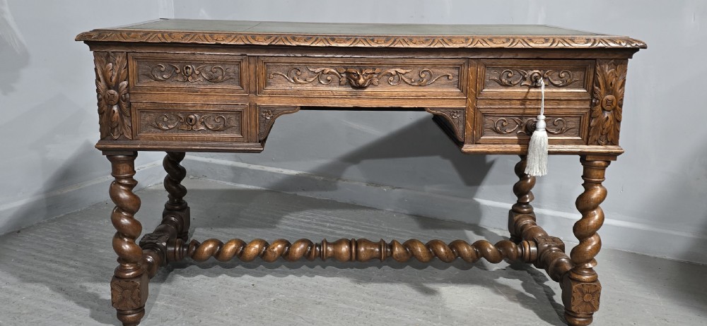 A carved oak writing desk featuring a green leather top insert, detailed scrollwork drawers with a central mask motif, and heavy barley-twist legs connected by a low stretcher. View III