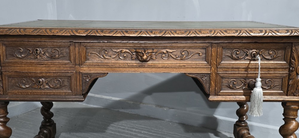A carved oak writing desk featuring a green leather top insert, detailed scrollwork drawers with a central mask motif, and heavy barley-twist legs connected by a low stretcher. View IV