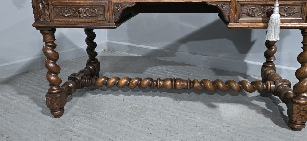 A carved oak writing desk featuring a green leather top insert, detailed scrollwork drawers with a central mask motif, and heavy barley-twist legs connected by a low stretcher. View V