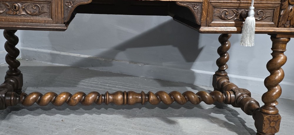 A carved oak writing desk featuring a green leather top insert, detailed scrollwork drawers with a central mask motif, and heavy barley-twist legs connected by a low stretcher. View VI