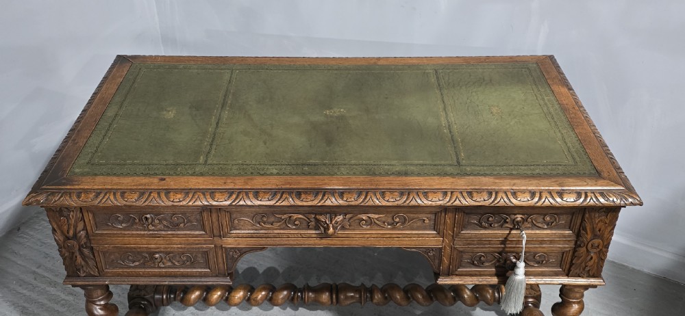 A carved oak writing desk featuring a green leather top insert, detailed scrollwork drawers with a central mask motif, and heavy barley-twist legs connected by a low stretcher. View VIII