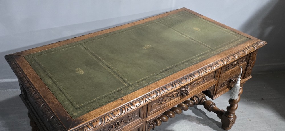 A carved oak writing desk featuring a green leather top insert, detailed scrollwork drawers with a central mask motif, and heavy barley-twist legs connected by a low stretcher. View IX