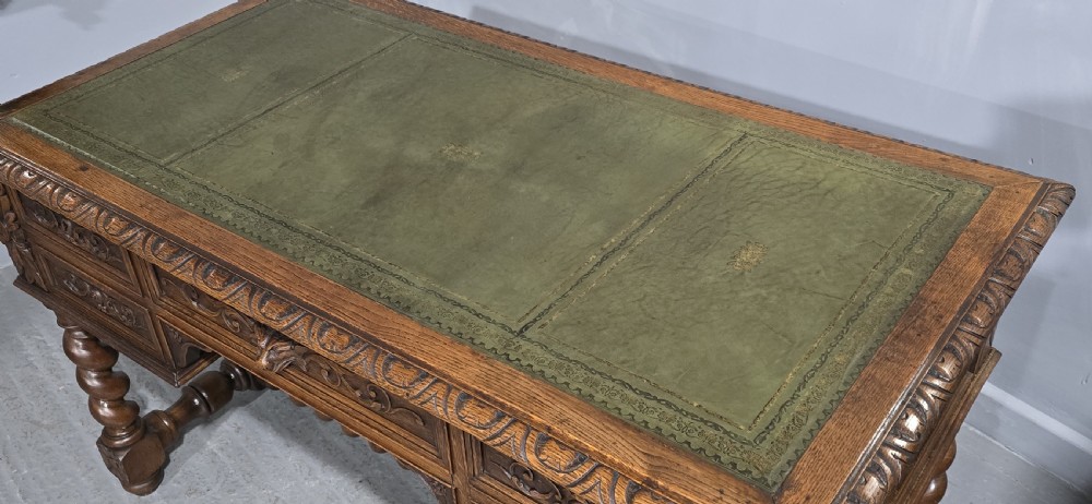 A carved oak writing desk featuring a green leather top insert, detailed scrollwork drawers with a central mask motif, and heavy barley-twist legs connected by a low stretcher. View X