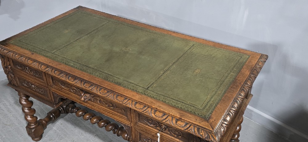 A carved oak writing desk featuring a green leather top insert, detailed scrollwork drawers with a central mask motif, and heavy barley-twist legs connected by a low stretcher. View XI