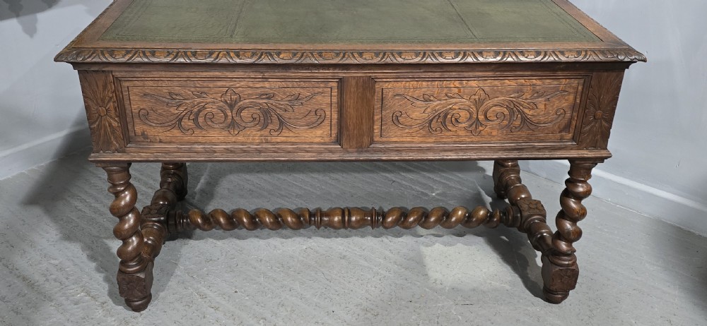 A carved oak writing desk featuring a green leather top insert, detailed scrollwork drawers with a central mask motif, and heavy barley-twist legs connected by a low stretcher. View XIII