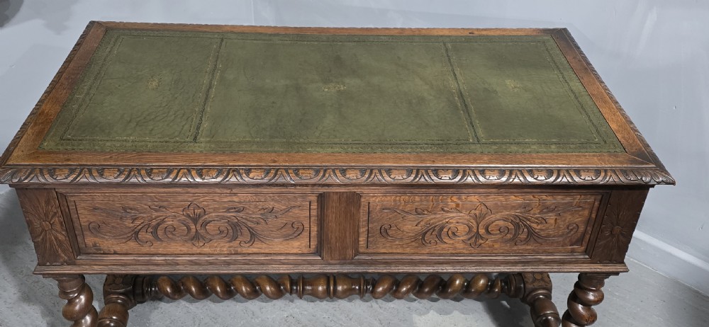 A carved oak writing desk featuring a green leather top insert, detailed scrollwork drawers with a central mask motif, and heavy barley-twist legs connected by a low stretcher. View XIV