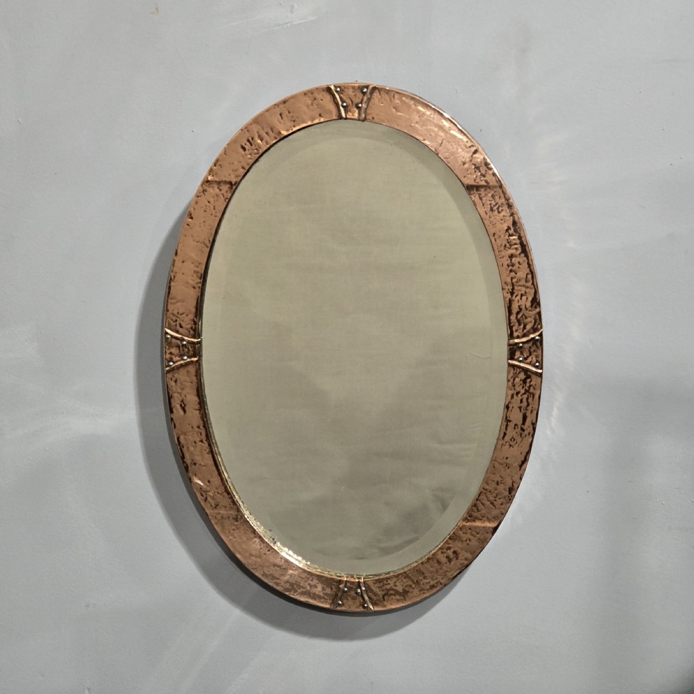 An oval wall mirror featuring a hand-hammered copper frame with raised riveted detail straps at four points and a bevelled glass plate.