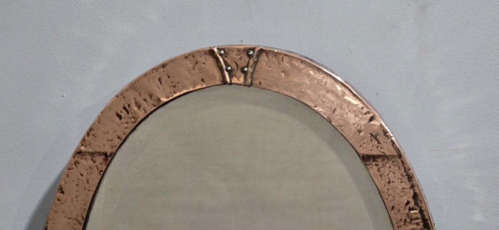An oval wall mirror featuring a hand-hammered copper frame with raised riveted detail straps at four points and a bevelled glass plate. View III