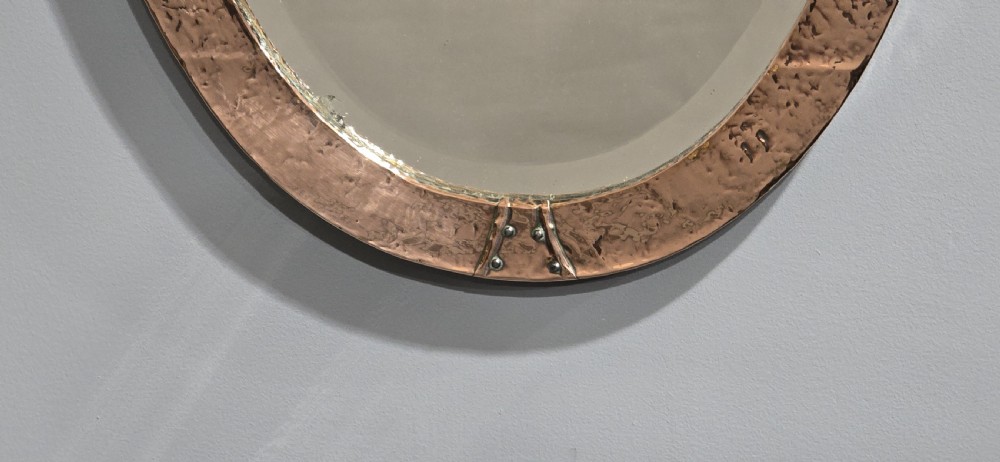 An oval wall mirror featuring a hand-hammered copper frame with raised riveted detail straps at four points and a bevelled glass plate. View V