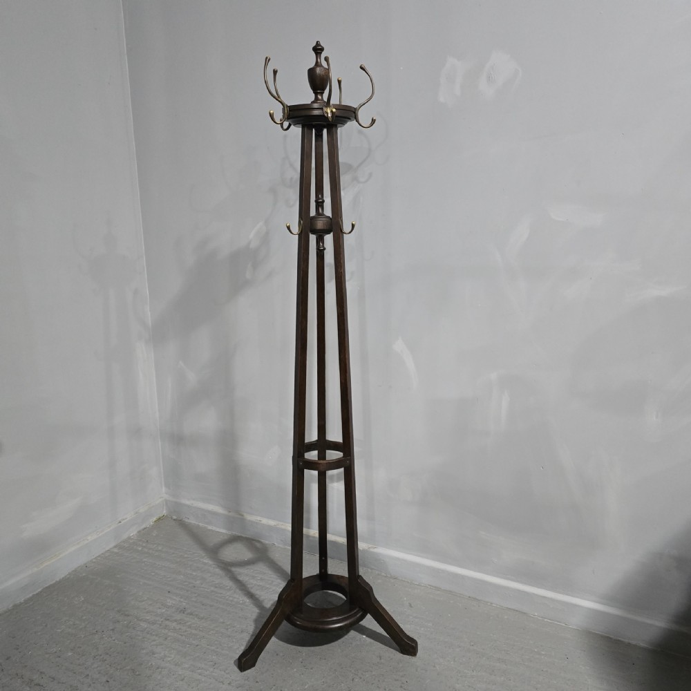 A tall dark oak Arts and Crafts freestanding coat stand featuring upper brass hooks, tapered uprights, a central umbrella ring, and splayed feet.
