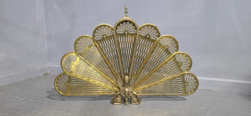 An antique brass peacock fire screen featuring openwork folding fan blades anchored by a cast winged creature pedestal. View II