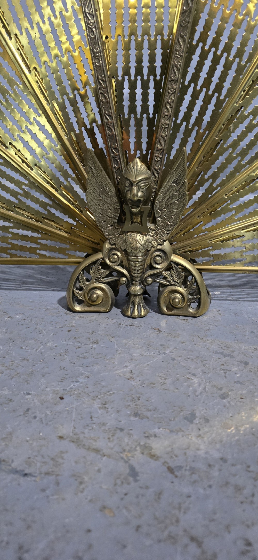 An antique brass peacock fire screen featuring openwork folding fan blades anchored by a cast winged creature pedestal. View III
