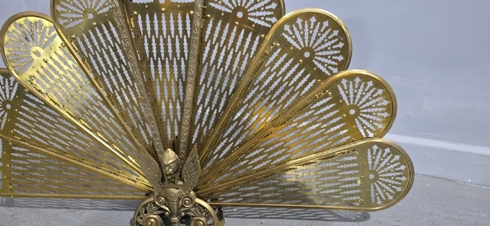 An antique brass peacock fire screen featuring openwork folding fan blades anchored by a cast winged creature pedestal. View IV