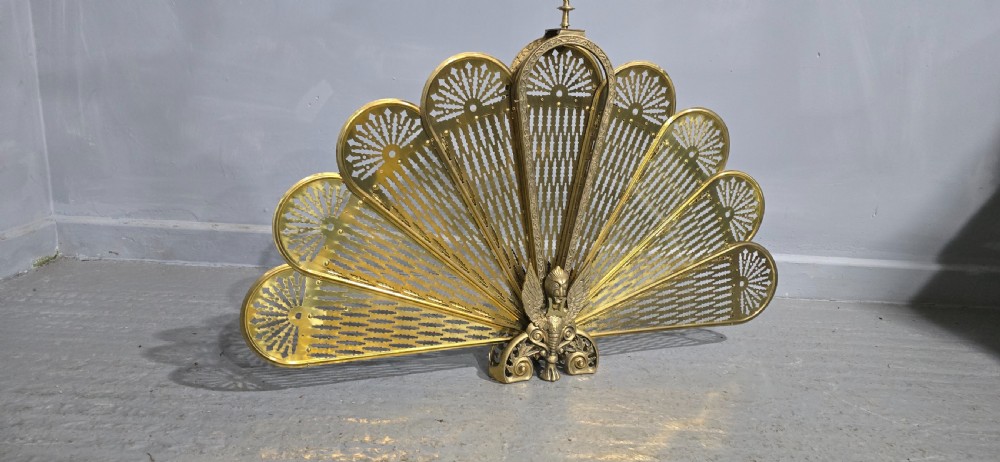 An antique brass peacock fire screen featuring openwork folding fan blades anchored by a cast winged creature pedestal. View V