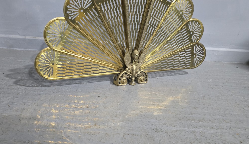 An antique brass peacock fire screen featuring openwork folding fan blades anchored by a cast winged creature pedestal. View VI