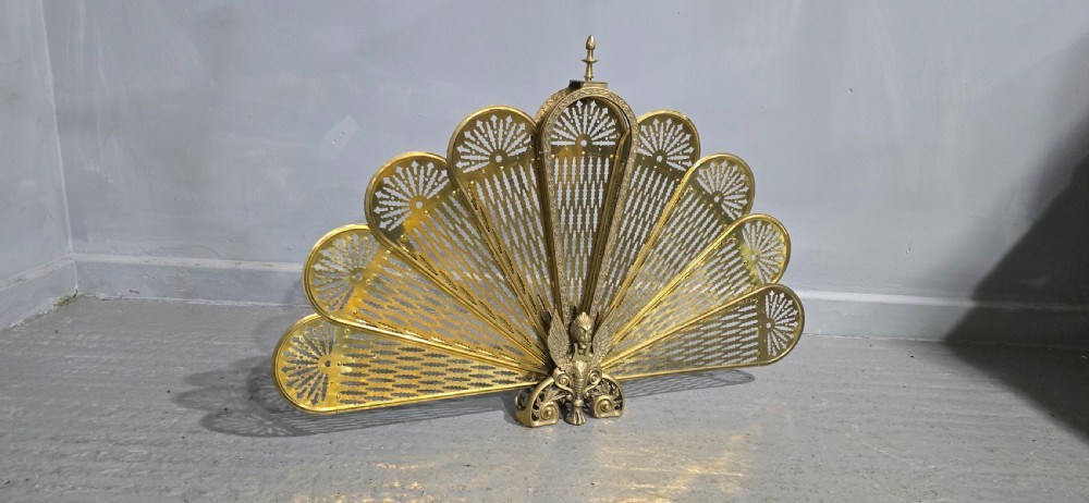 An antique brass peacock fire screen featuring openwork folding fan blades anchored by a cast winged creature pedestal. View VII