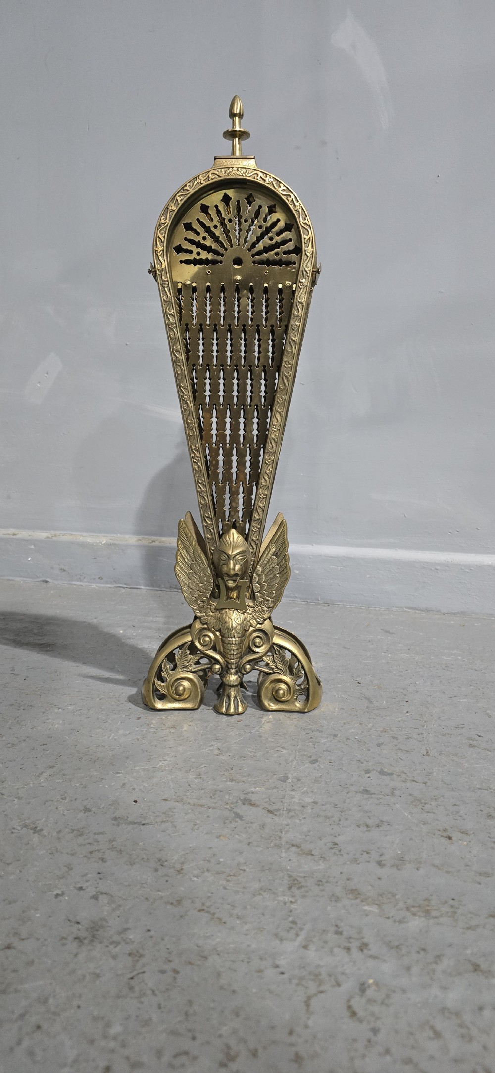 An antique brass peacock fire screen featuring openwork folding fan blades anchored by a cast winged creature pedestal. View VIII