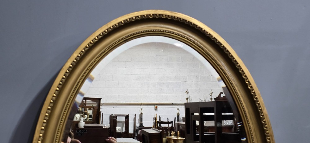 An oval wall mirror framed in a gold gilt moulded rim with beaded detailing. View VI