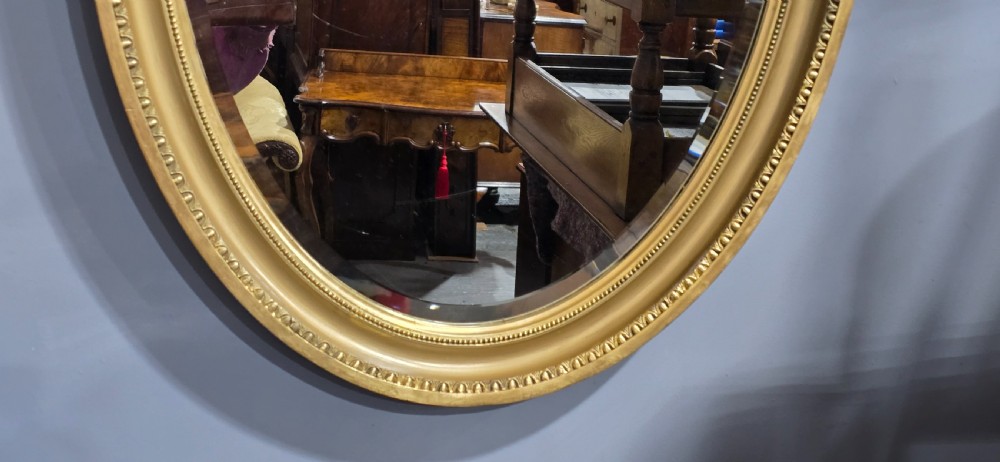 An oval wall mirror framed in a gold gilt moulded rim with beaded detailing. View VII