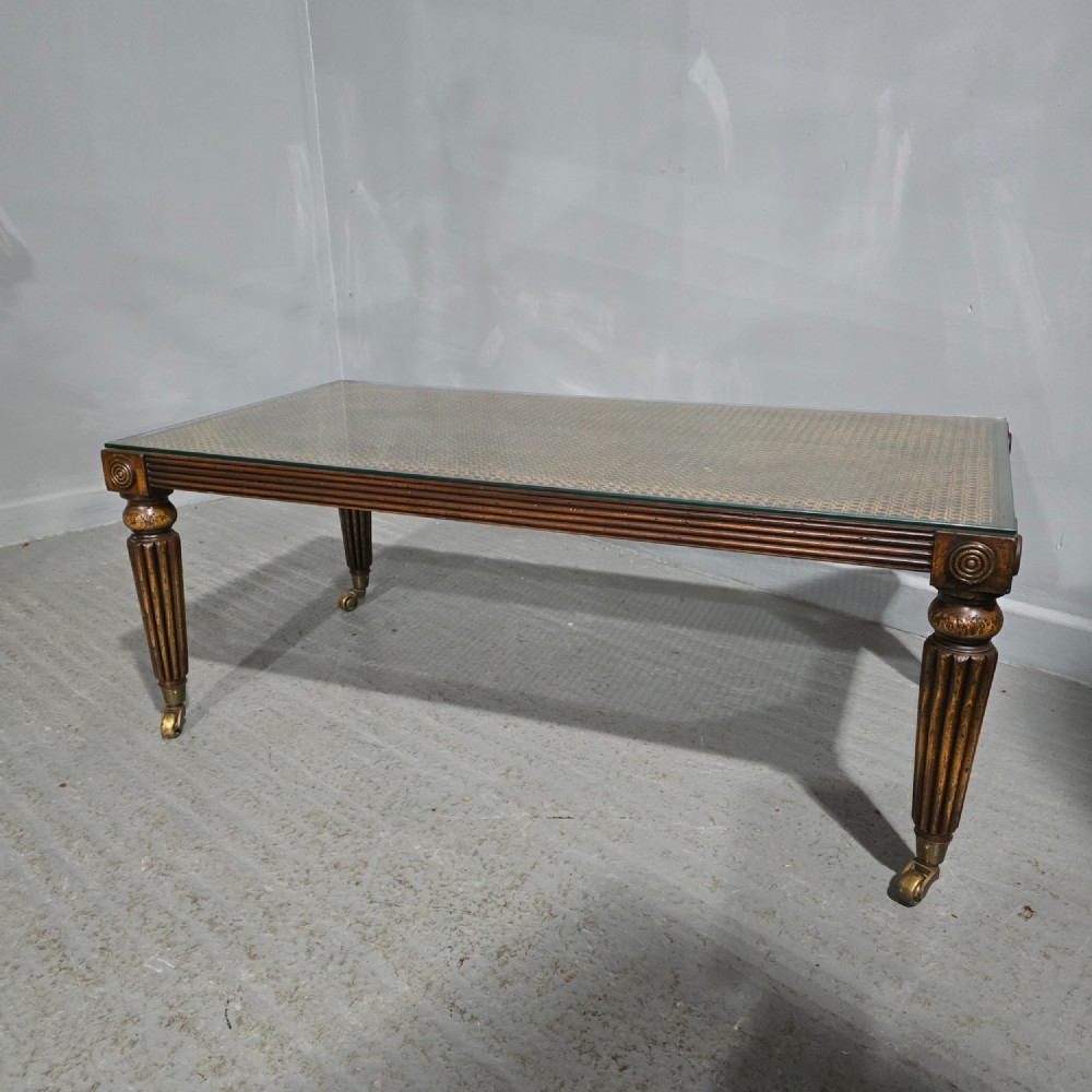 A low rectangular mahogany coffee table featuring a glass-covered woven cane top, reeded frieze, carved corner rosettes, and tapered reeded legs terminating in brass castors.