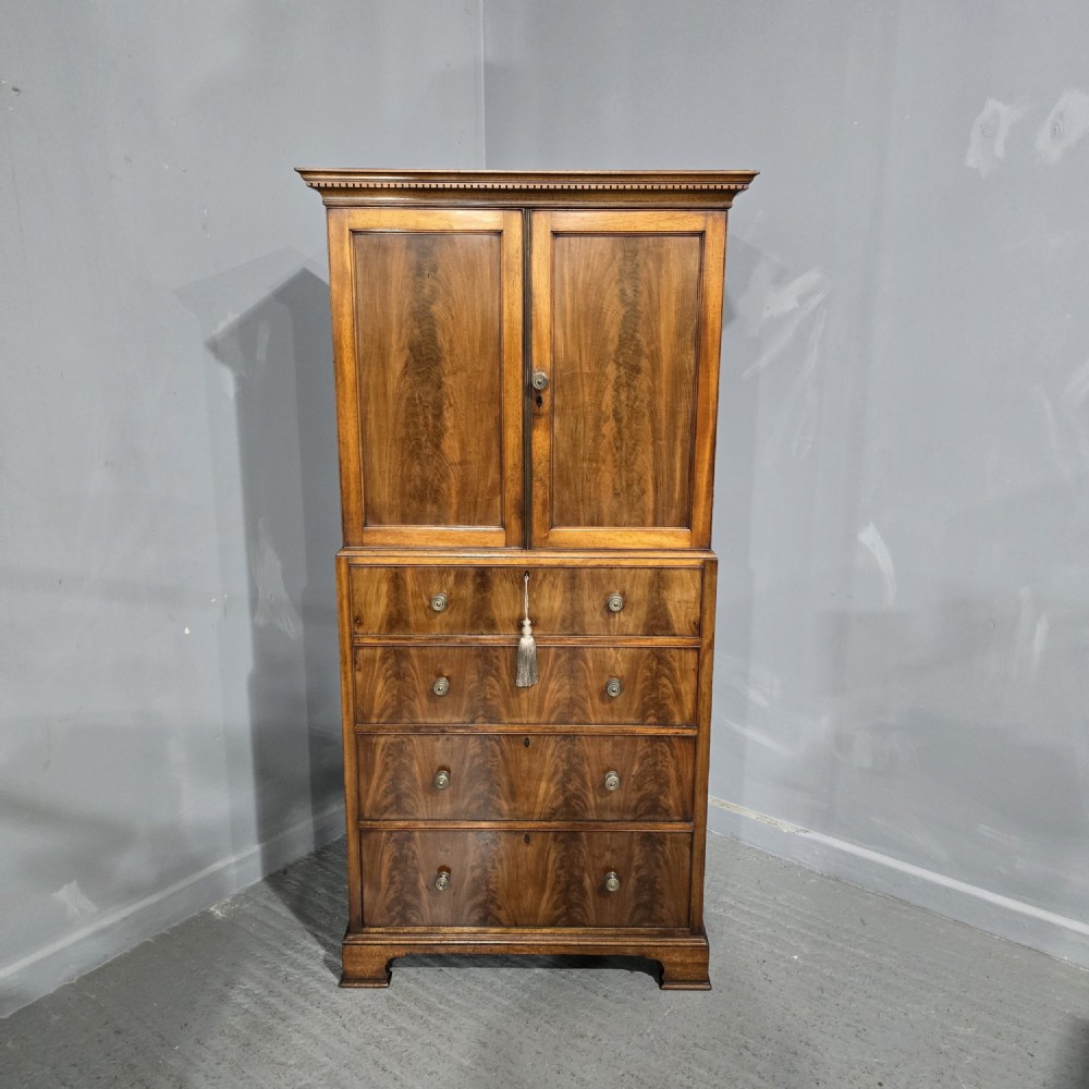 A tall mahogany linen press featuring a double-door upper cupboard with flame mahogany panels over four long drawers with turned knobs.