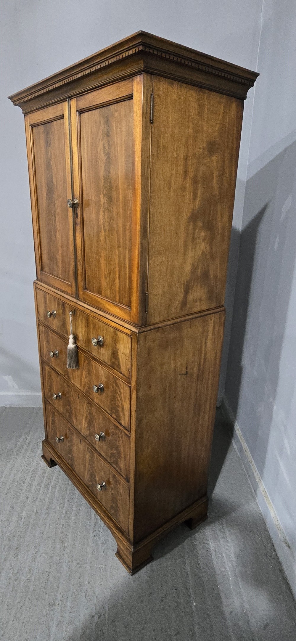 A tall mahogany linen press featuring a double-door upper cupboard with flame mahogany panels over four long drawers with turned knobs. View VI