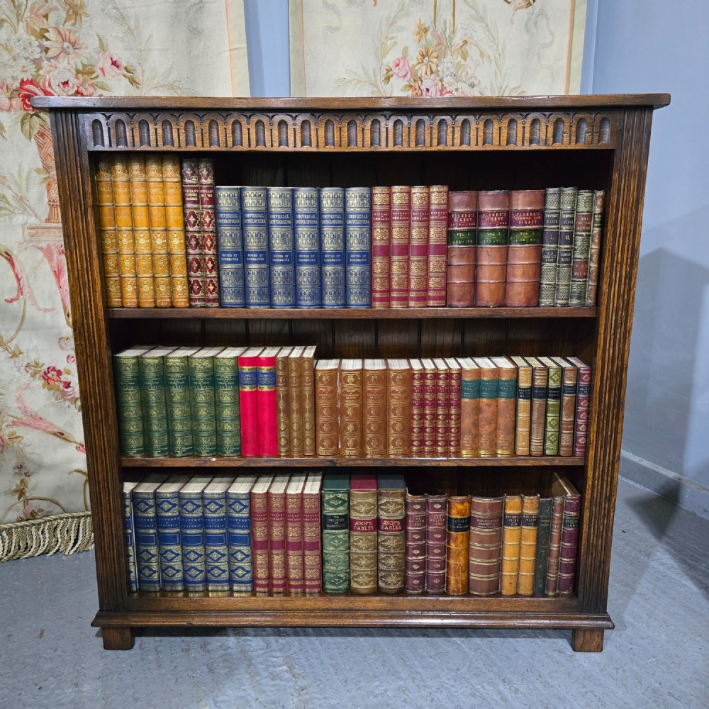 A low solid oak open bookcase with three shelves filled with antiquarian books, featuring a carved arcaded frieze and reeded side moulding.