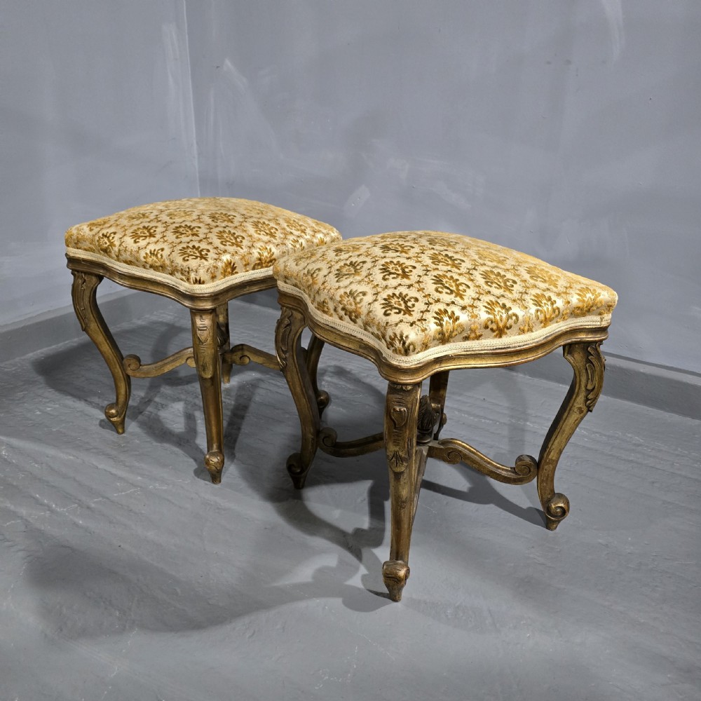 A pair of French giltwood stools featuring patterned gold velvet upholstery, carved cabriole legs, and ornate stretchers.