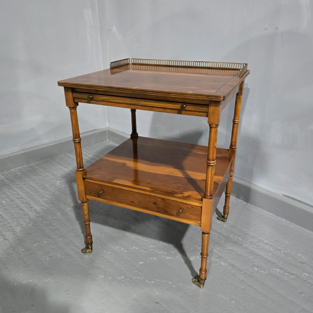 A two-tiered yew wood lamp table featuring a brass gallery top rail, turned legs, a lower drawer, and brass castors.