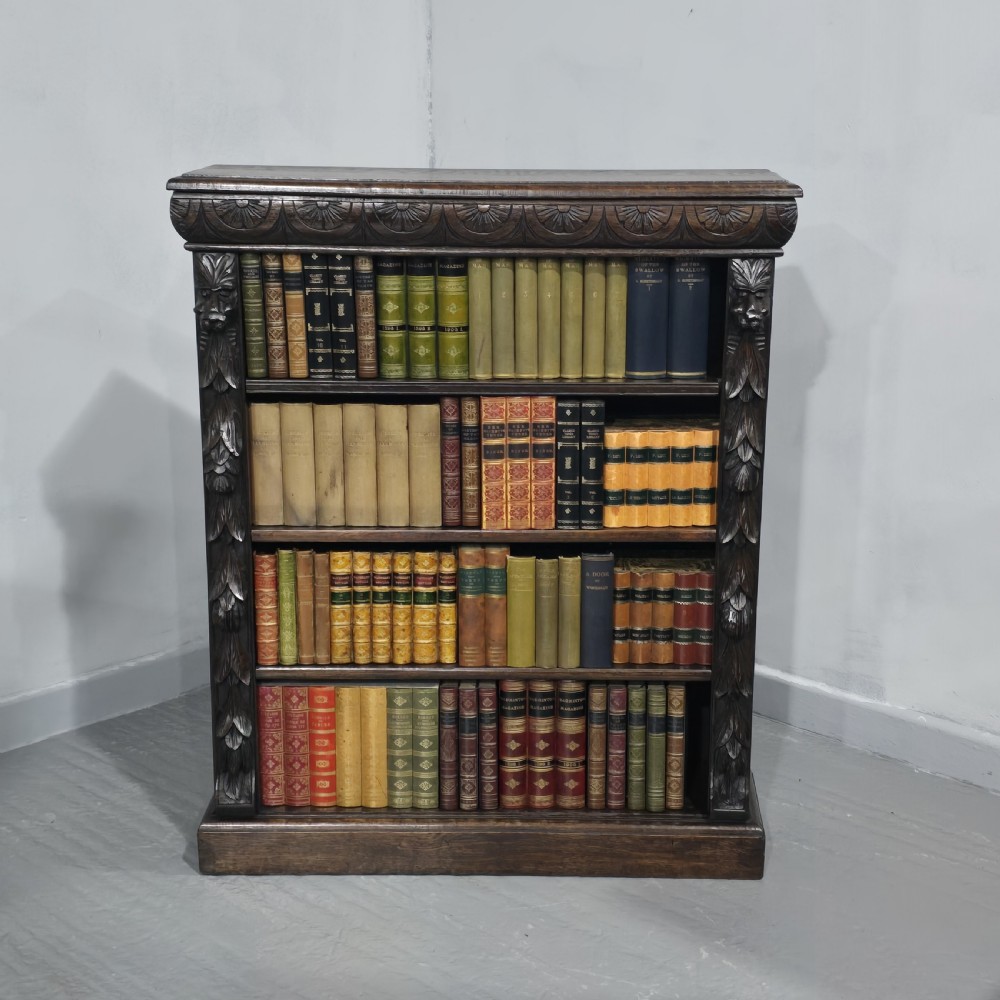 A dark carved oak open bookcase featuring four shelves filled with leather-bound books, flanked by carved foliate pilasters and topped with a decorative frieze.