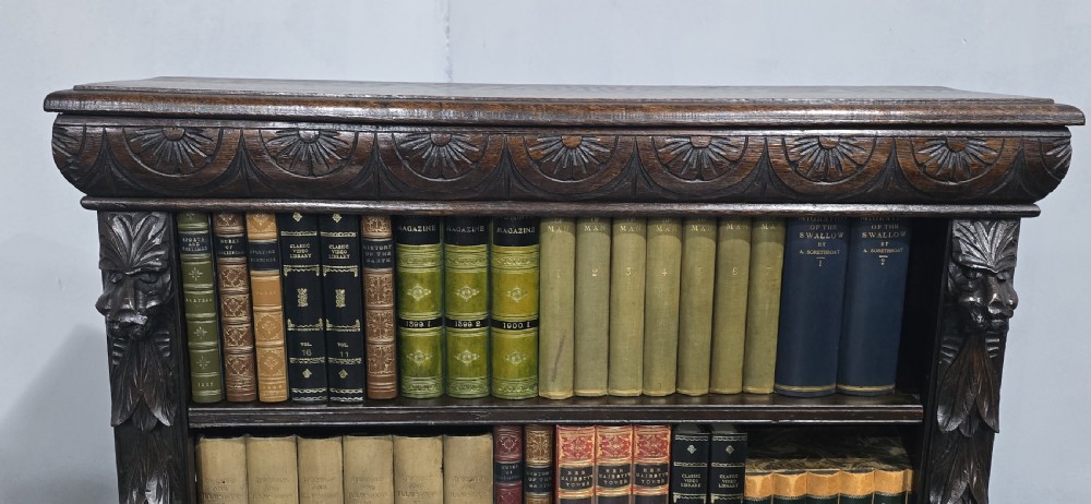 A dark carved oak open bookcase featuring four shelves filled with leather-bound books, flanked by carved foliate pilasters and topped with a decorative frieze. View VI